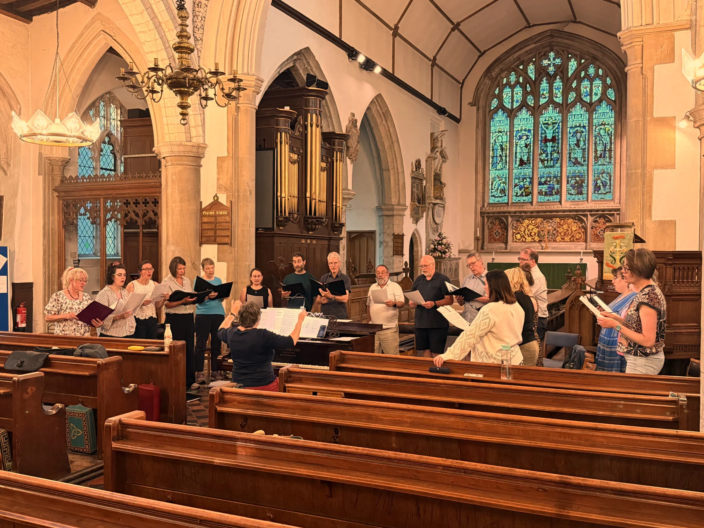 The choir standing in a line in All Saints' Church, Writtle, singing from their copies at a Monday evening rehearsal, with the conductor at the piano